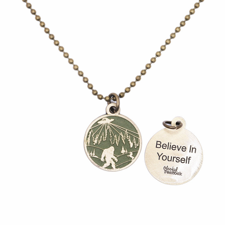 Whimsical charm with illustration of bigfoot, a UFO and a unicorn. The reverse says "Believe in Yourself." Antique bronze painted olive green and burnished.