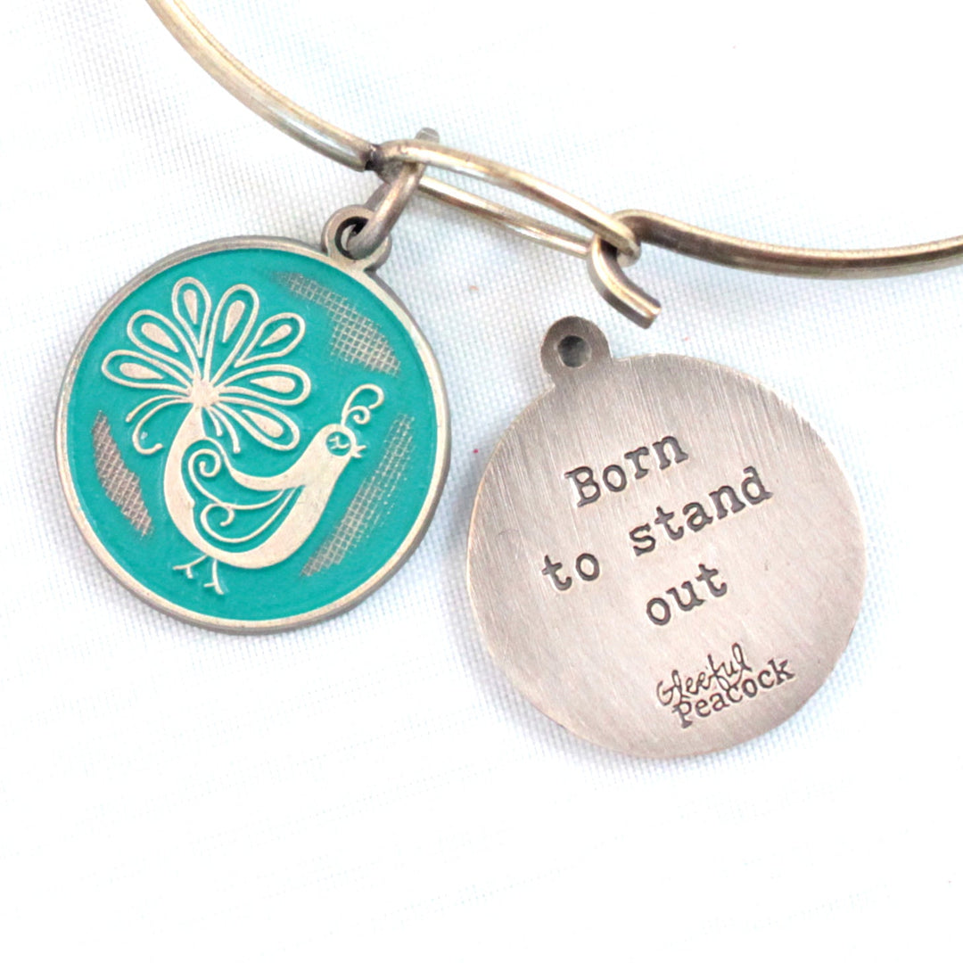 Antique bronze charm painted teal and burnished to shiny gold. One side depicts a whimsical peacock. Reverse side says "Born to stand out".