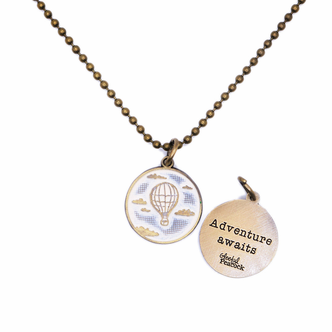 Antique bronze charm painted cream and burnished to shiny gold. One side depicts a hot air balloon floating in the clouds. Reverse side says "Adventure Awaits".