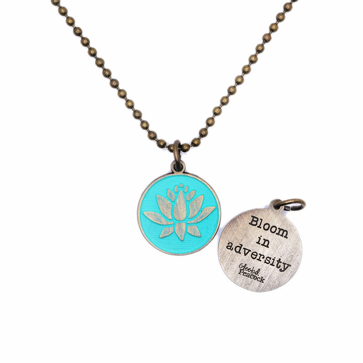 Antique bronze charm painted turquoise and burnished to shiny gold. One side depicts a blooming lotus flower. Reverse side says "Bloom in Adversity".