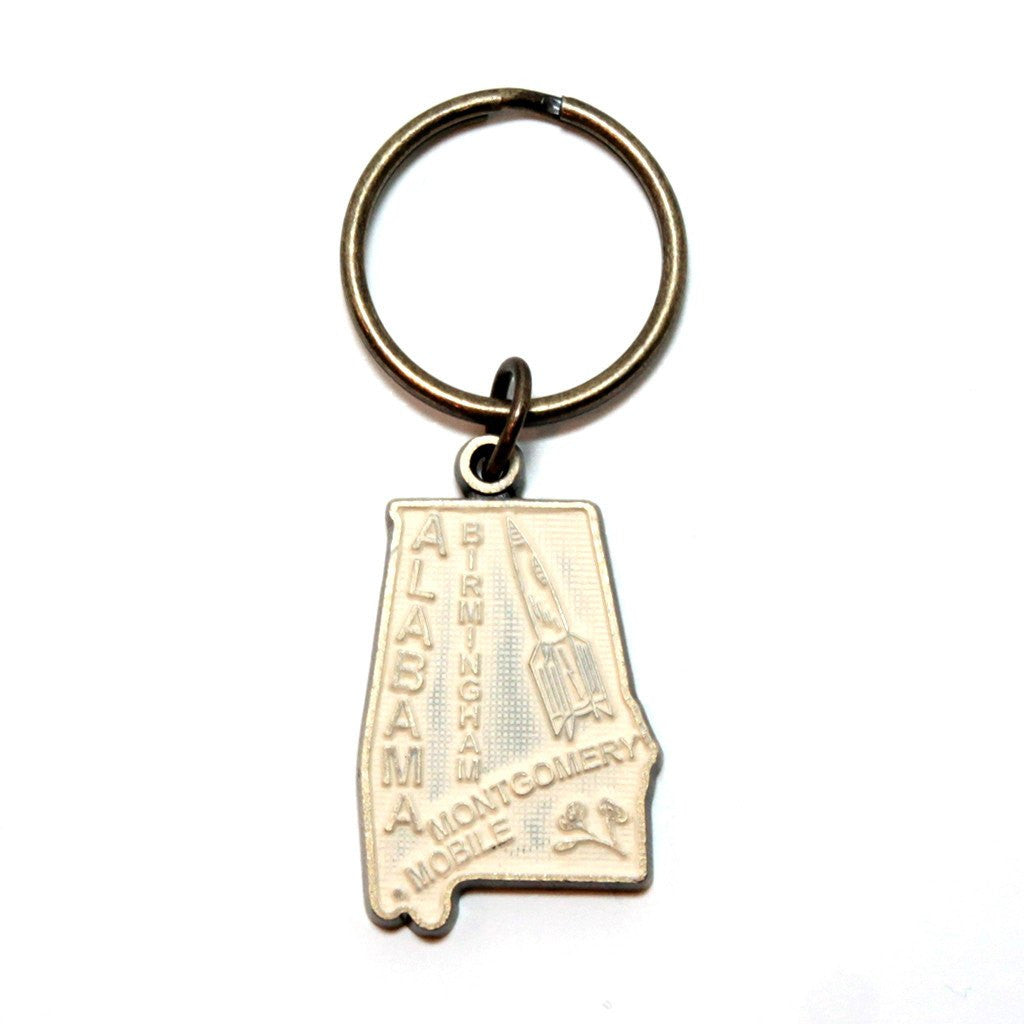 Antique bronze 1 1/4" charm keychain on 1 1/4" keyring. Alabama state charm painted cream and burnished to shiny gold. Map depicts location of Birmingham, Montgomery and Mobile and shows a rocket ship.