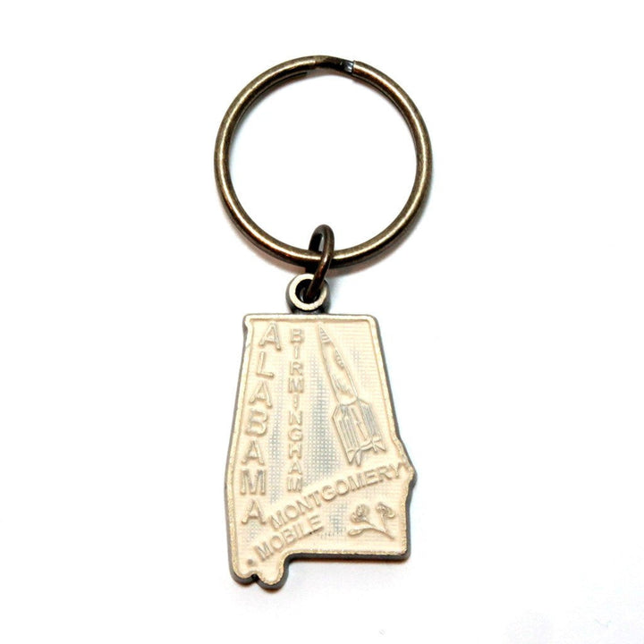 Antique bronze 1 1/4" charm keychain on 1 1/4" keyring. Alabama state charm painted cream and burnished to shiny gold. Map depicts location of Birmingham, Montgomery and Mobile and shows a rocket ship.